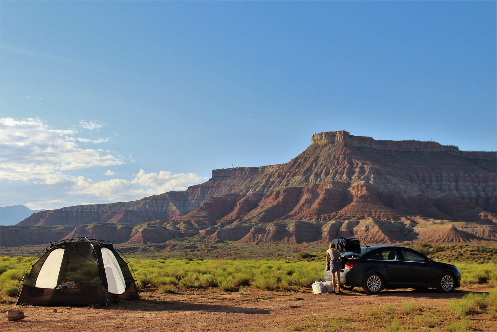 A tent and a small car parked in the desert, a woman getting something out of the trunk of the car