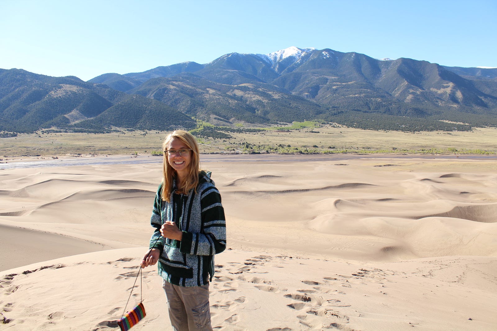 a young lady holding a small purse, standing on sand dunes with a mountain range behind her